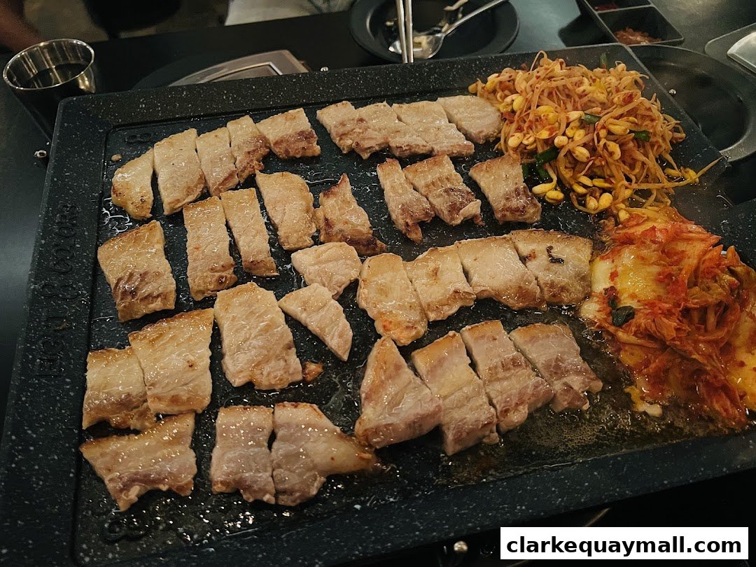 Slices of grilled pork belly with kimchi and bean sprouts on a hot plate.
