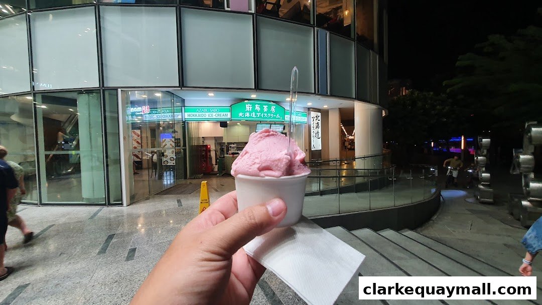 A hand holds a cup of pink ice cream in front of the Azabu Sabo Hokkaido Ice-Cream shop.