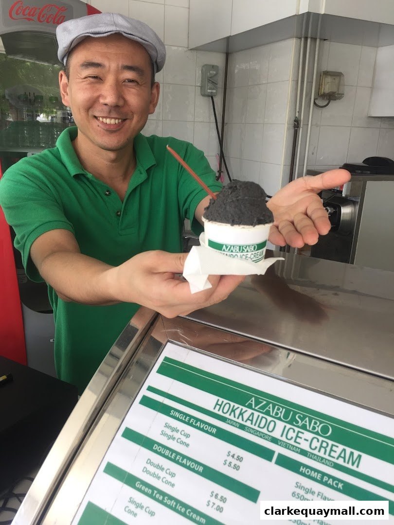 A smiling man in a green shirt and cap presents a cup of black ice cream.