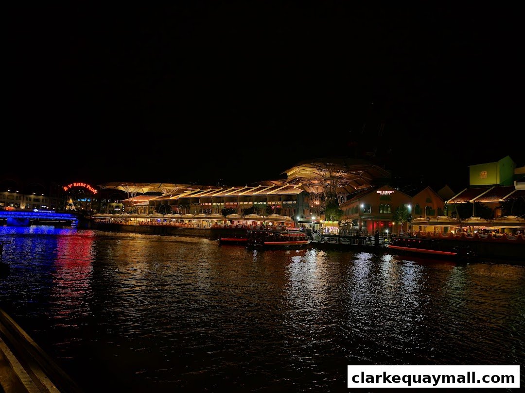 A scenic night view of Clarke Quay with illuminated buildings and boats along the Singapore River.