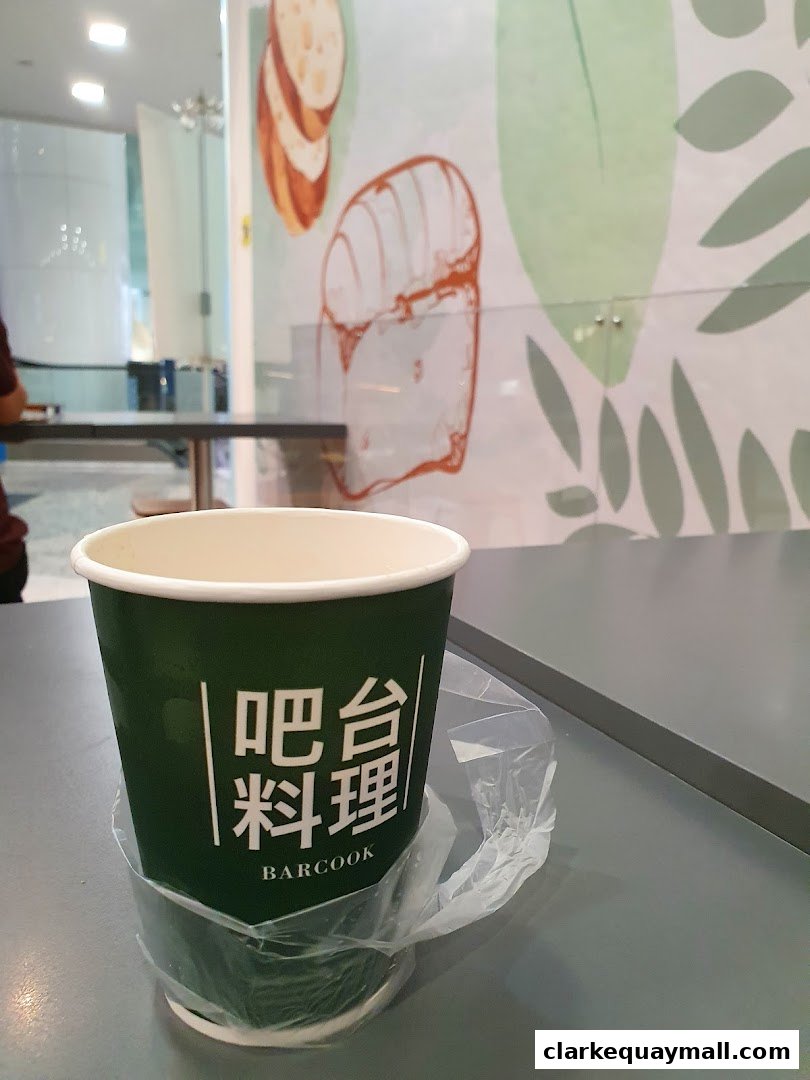 A green paper cup with the Barcook Bakery logo sits on a table with a plastic bag.