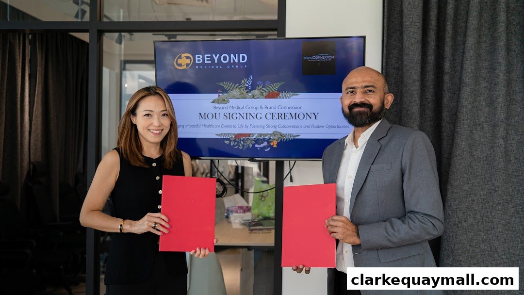 Two people smile while holding red folders in front of a screen displaying an MOU signing ceremony.