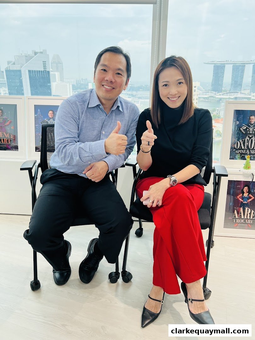 Two smiling individuals give thumbs up in an office with a city skyline view.