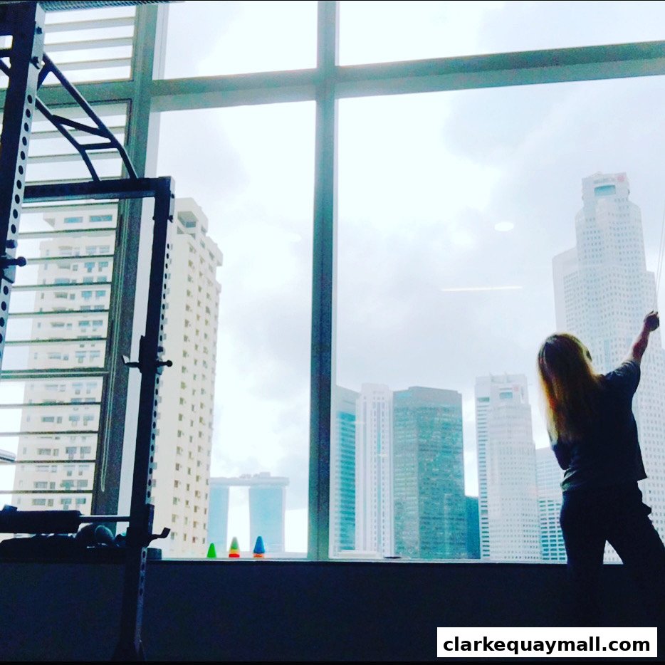 A person stands by a large window, looking out at a city skyline with gym equipment in the foreground.