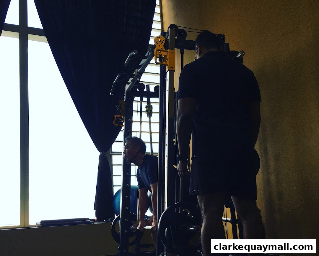 Two men are working out with weights in a gym, with a large window and gym equipment visible.