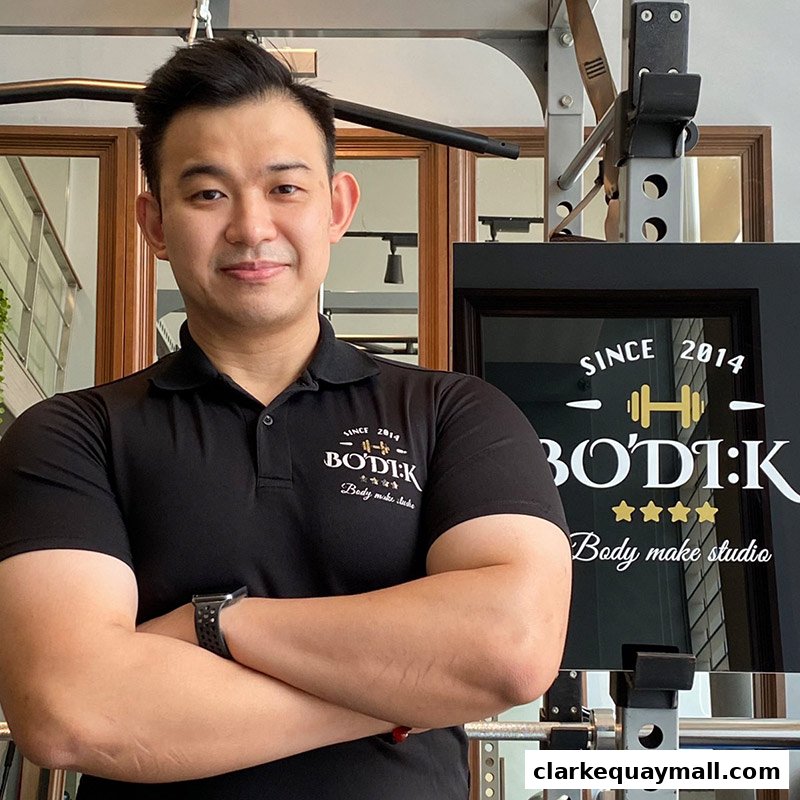 A man in a black polo shirt with the BODIK logo stands with his arms crossed in a gym.