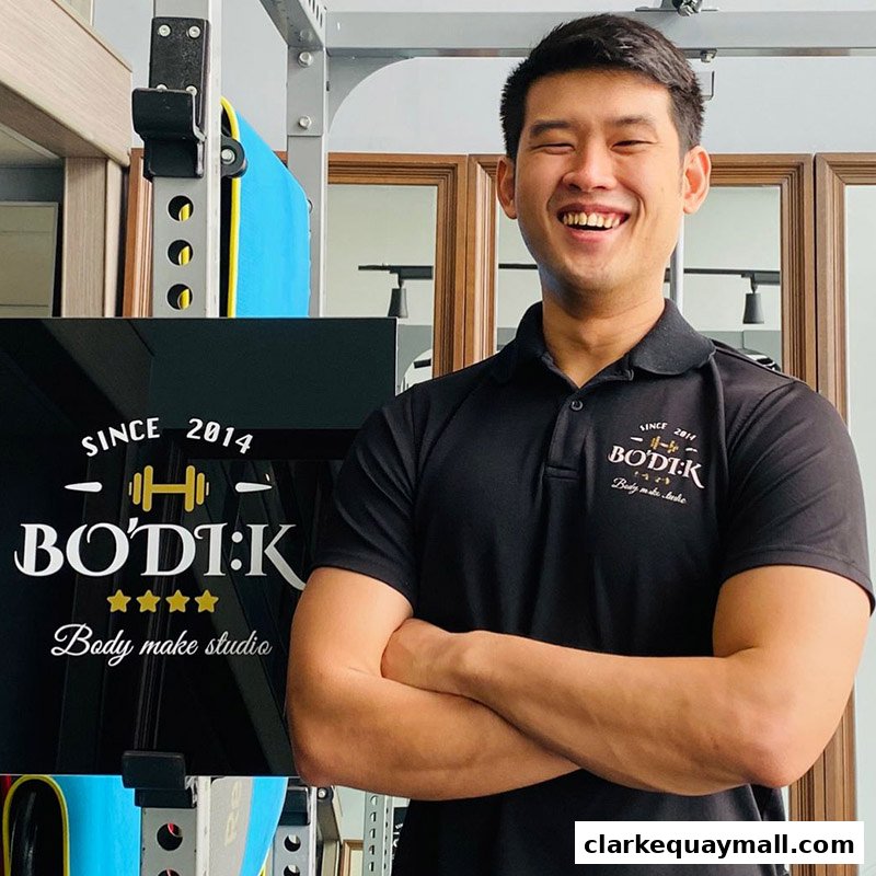 A smiling man in a black polo shirt stands with his arms crossed in front of a gym.