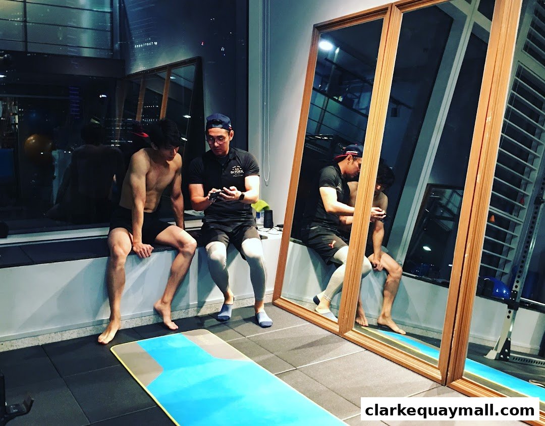 Two men are sitting in a gym, one is shirtless and the other is looking at his phone.