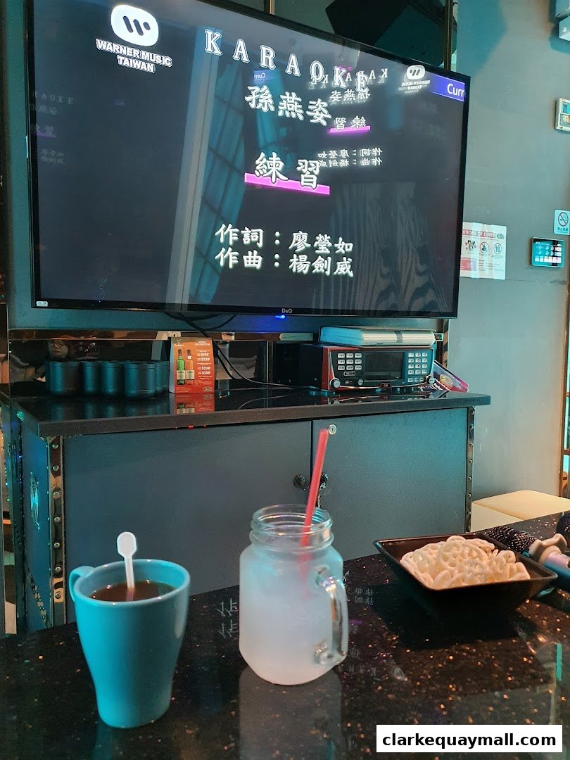 A screen displays a karaoke song with drinks and snacks on a counter.