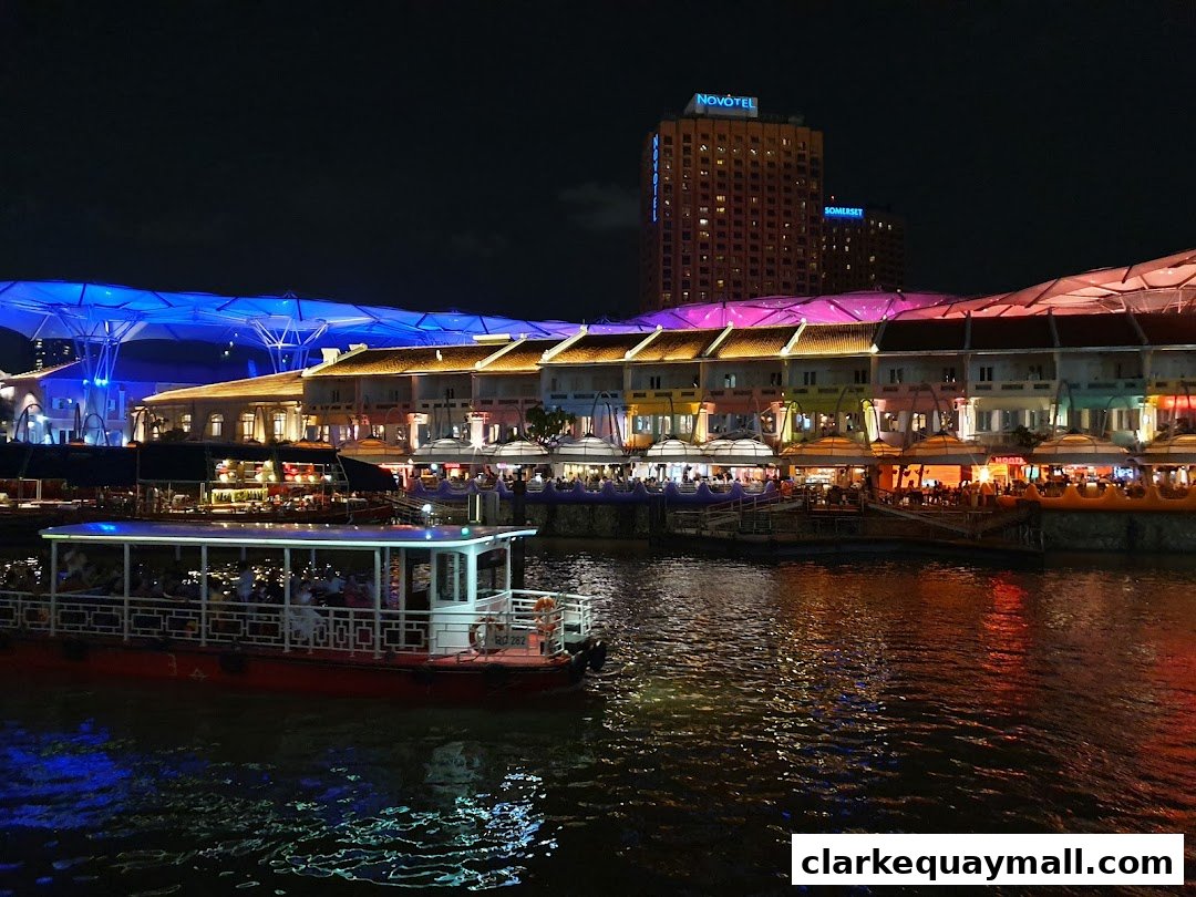 A scenic night view of Clarke Quay Central with illuminated buildings and a boat on the river.