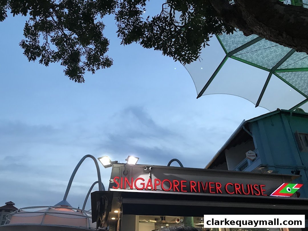 A sign for Singapore River Cruise is illuminated against a twilight sky with trees and a modern canopy.