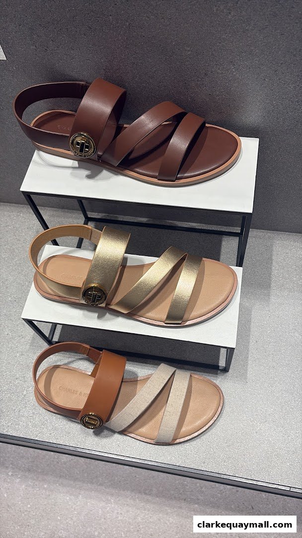 Three stylish flat sandals in brown, gold, and tan/brown displayed on white shelves.