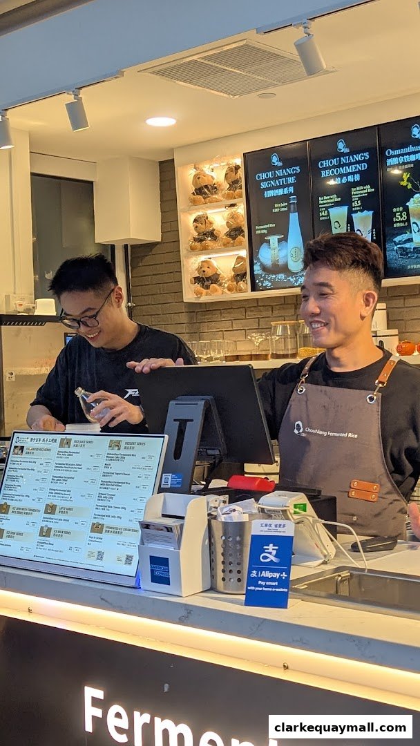 Two staff members behind a counter at Chou Niang Fermented Rice, with a menu and payment options displayed.