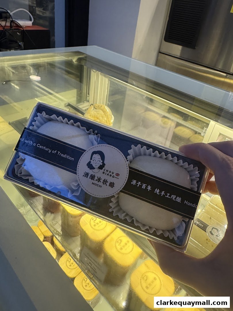 A hand holds a box of mochi in front of a display case filled with desserts.