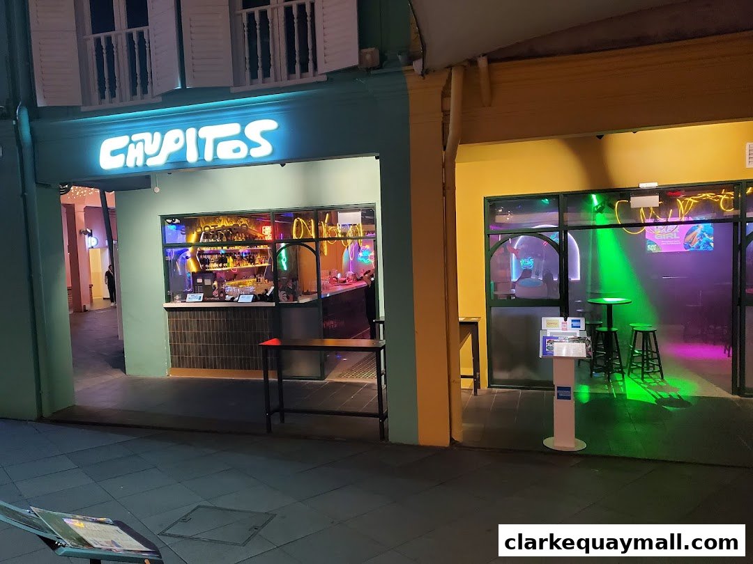 The exterior of Chupitos, a bar with a neon sign and colorful interior lighting.