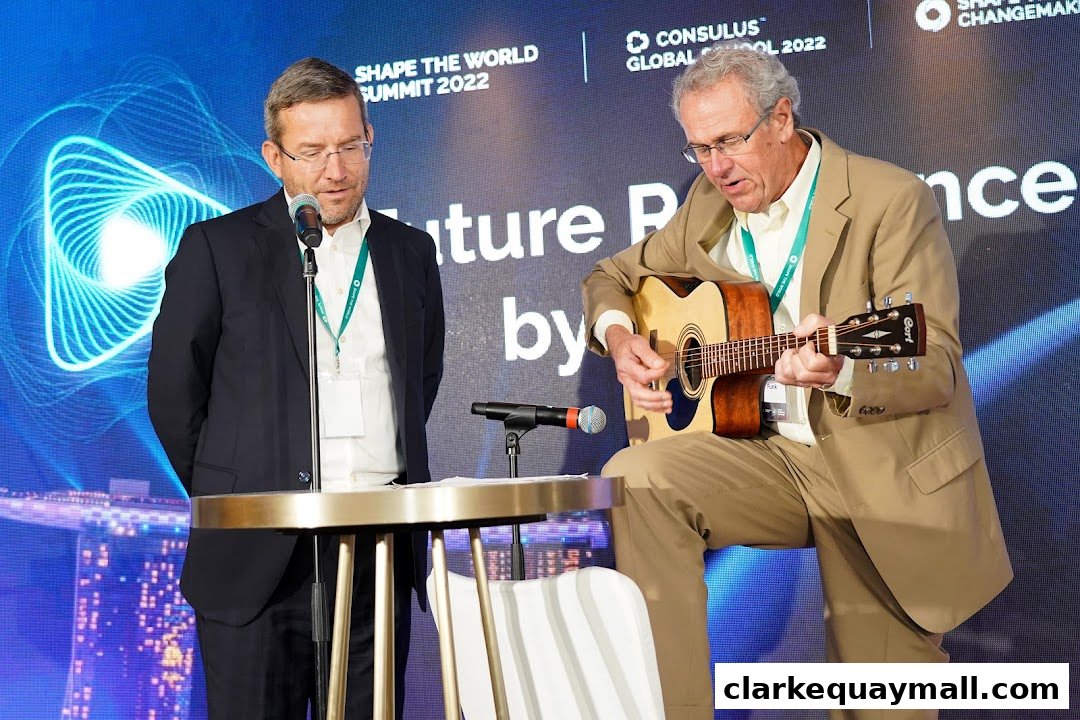 Two men on stage, one speaking into a microphone, the other playing an acoustic guitar.