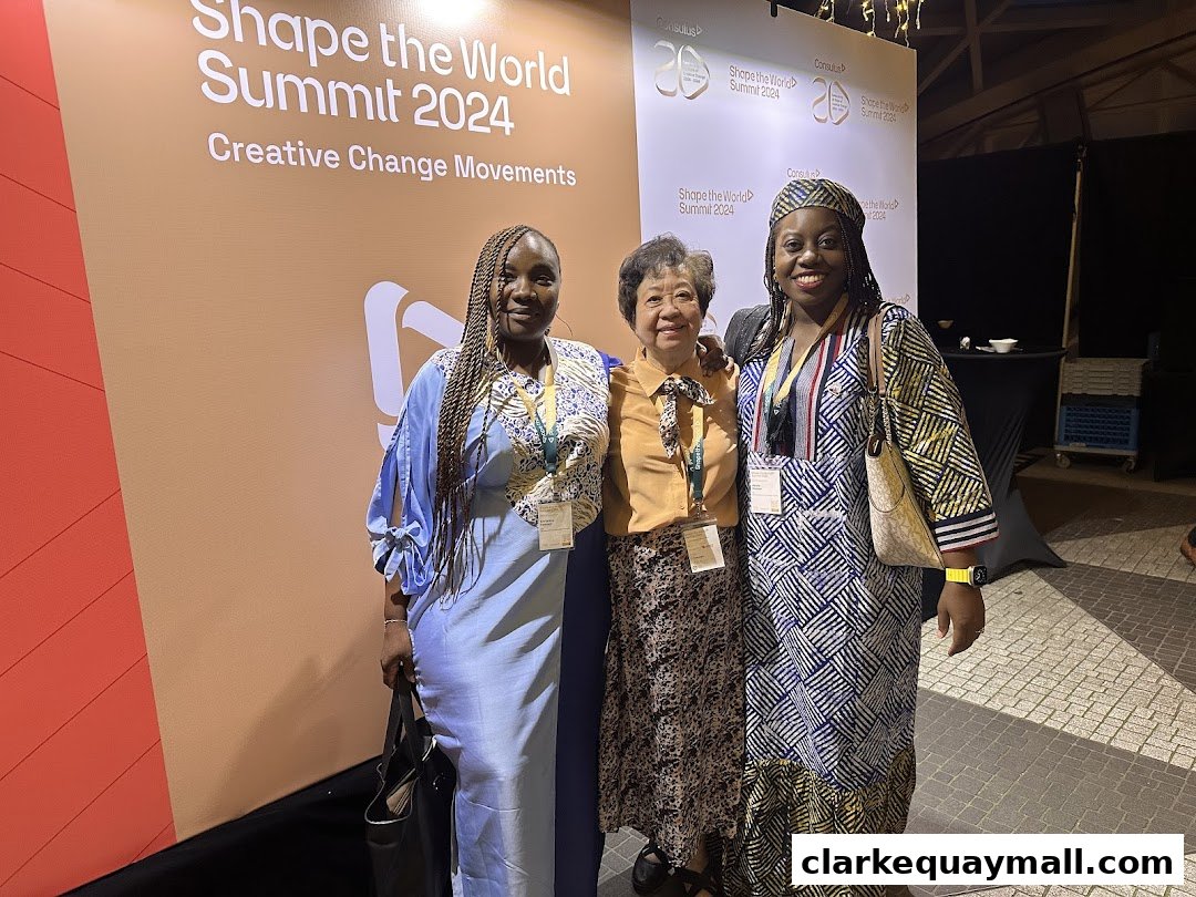 Three women pose in front of a backdrop for the 'Shape the World Summit 2024'.