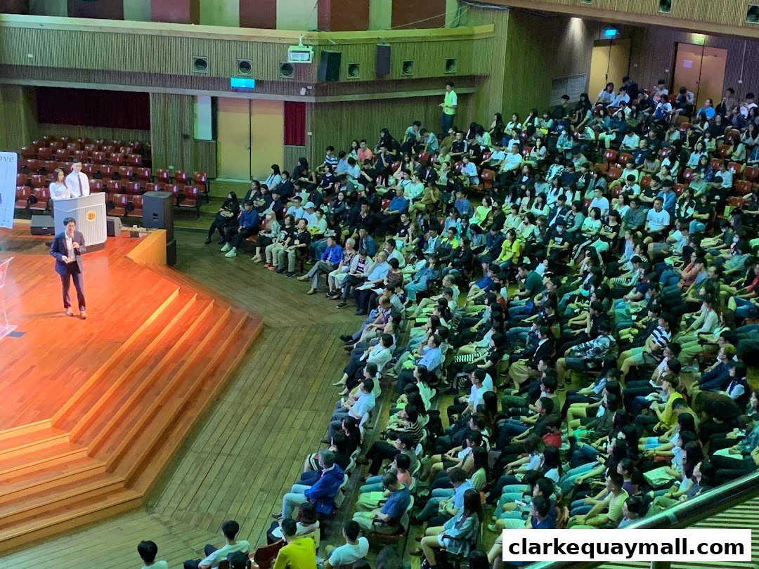 A speaker addresses a large audience seated in an auditorium with tiered seating.