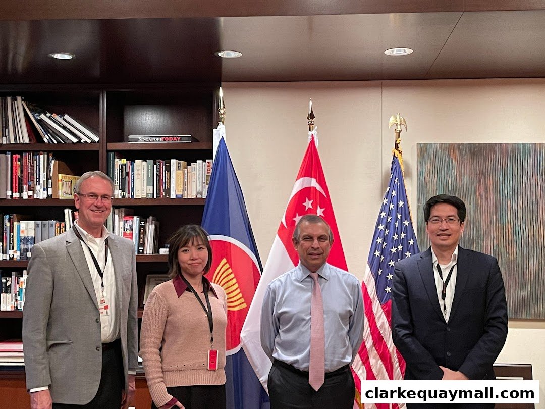 Four individuals stand in front of bookshelves and flags, including Singapore and the USA.