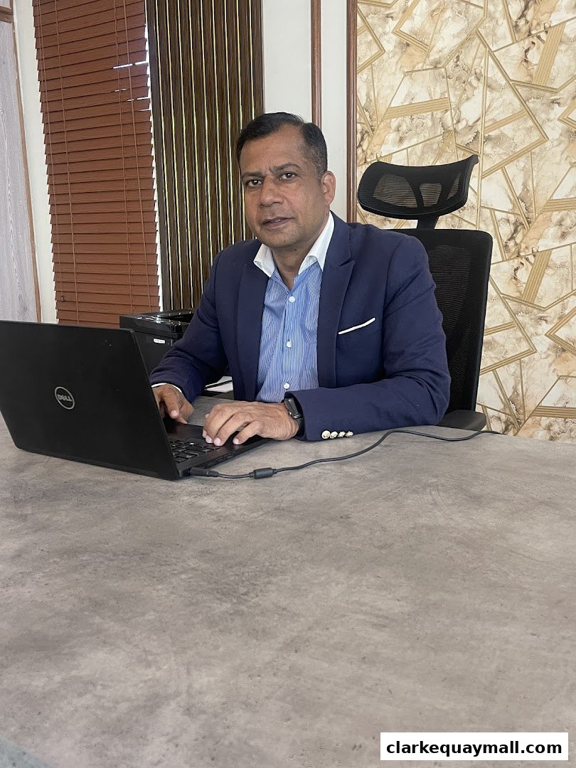 A man in a suit sits at a desk with a laptop, looking towards the camera.