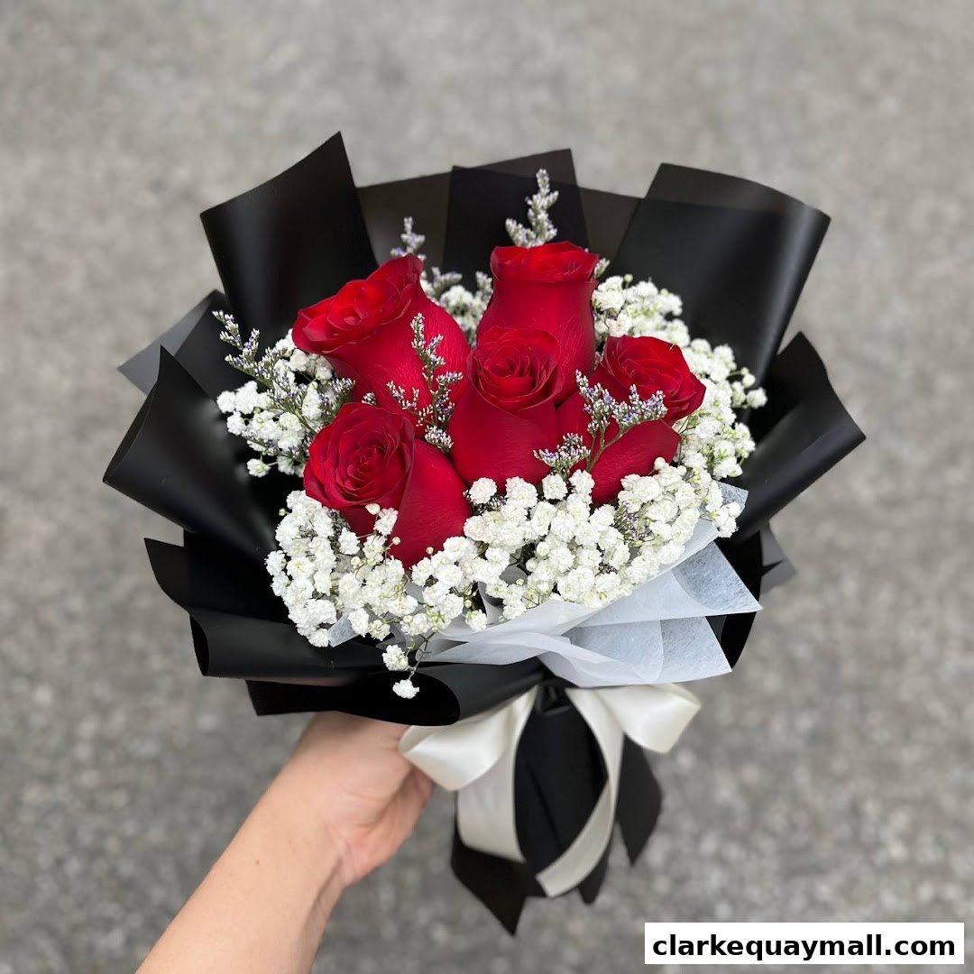A hand holds a bouquet of red roses with baby's breath, wrapped in black paper.