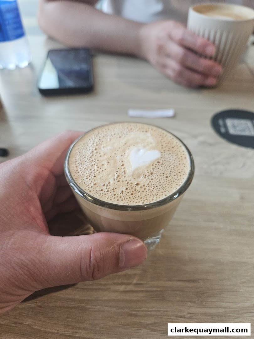A hand holds a glass of coffee with latte art, with other drinks and a phone on the table.