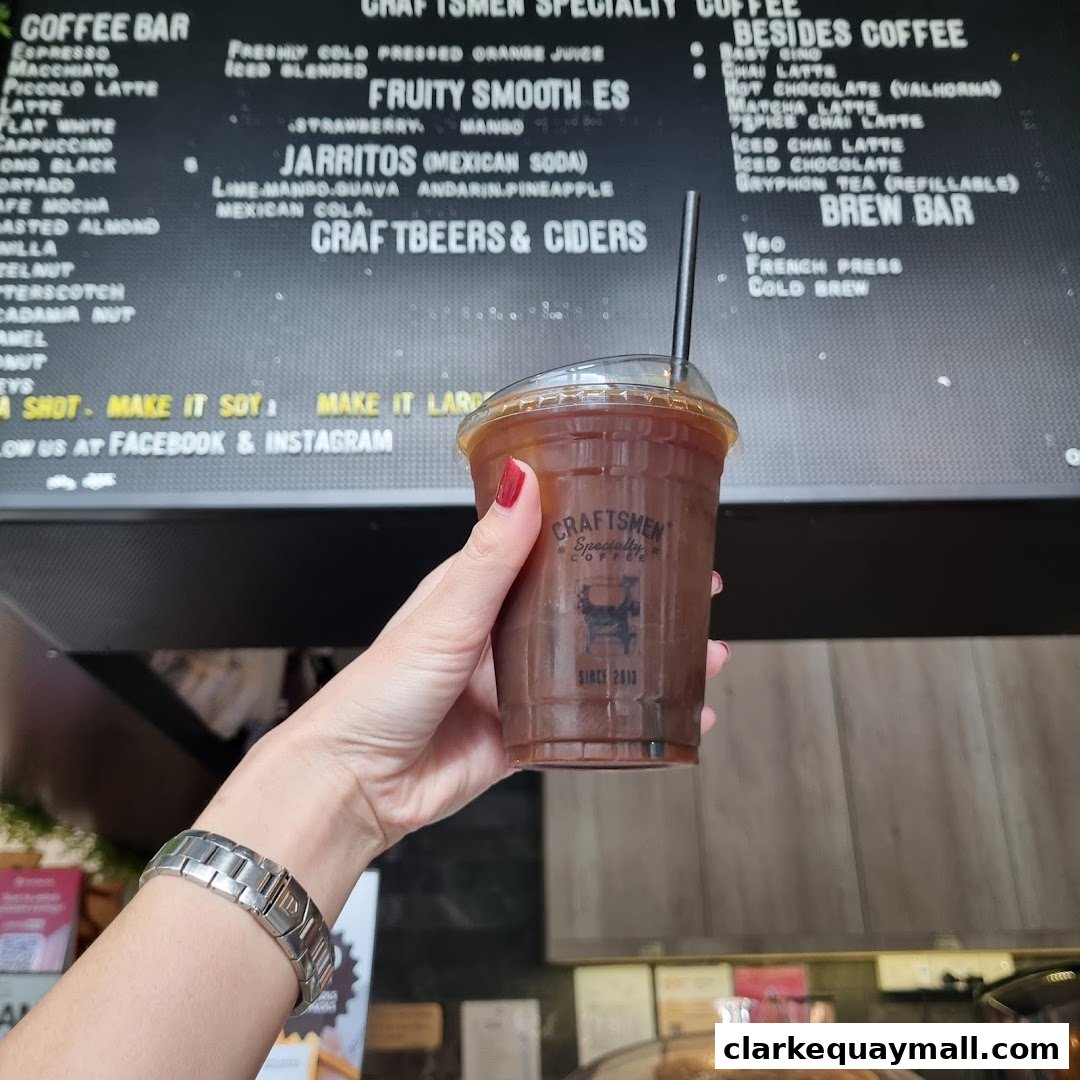A hand holds a cold coffee drink in front of a menu board at Craftsmen Coffee.