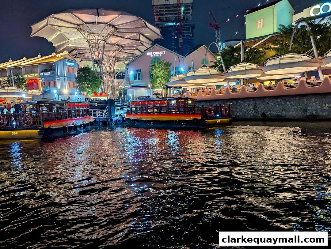 Night view of boats docked on a river with illuminated buildings and a distinctive canopy structure.