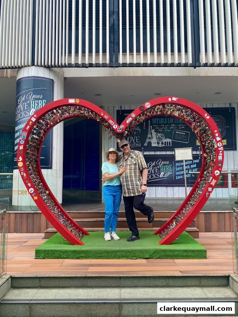 A couple poses in front of a large red heart sculpture adorned with love locks.