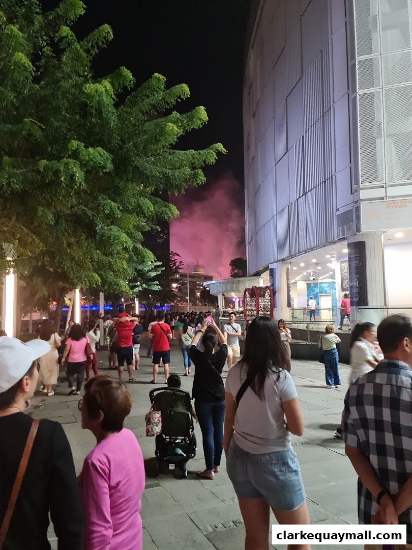People gather on a street at night, with a tree and a building in the background.