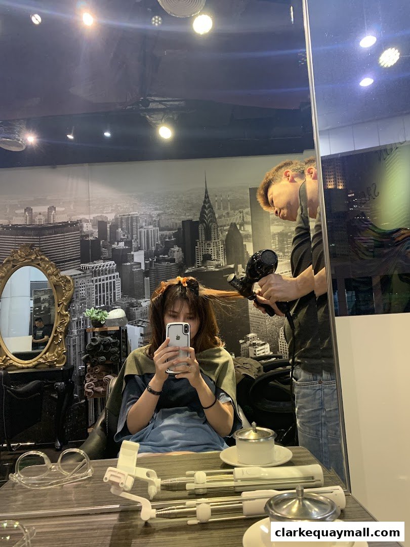 A stylist blow-dries a client's hair while she takes a selfie in a salon.