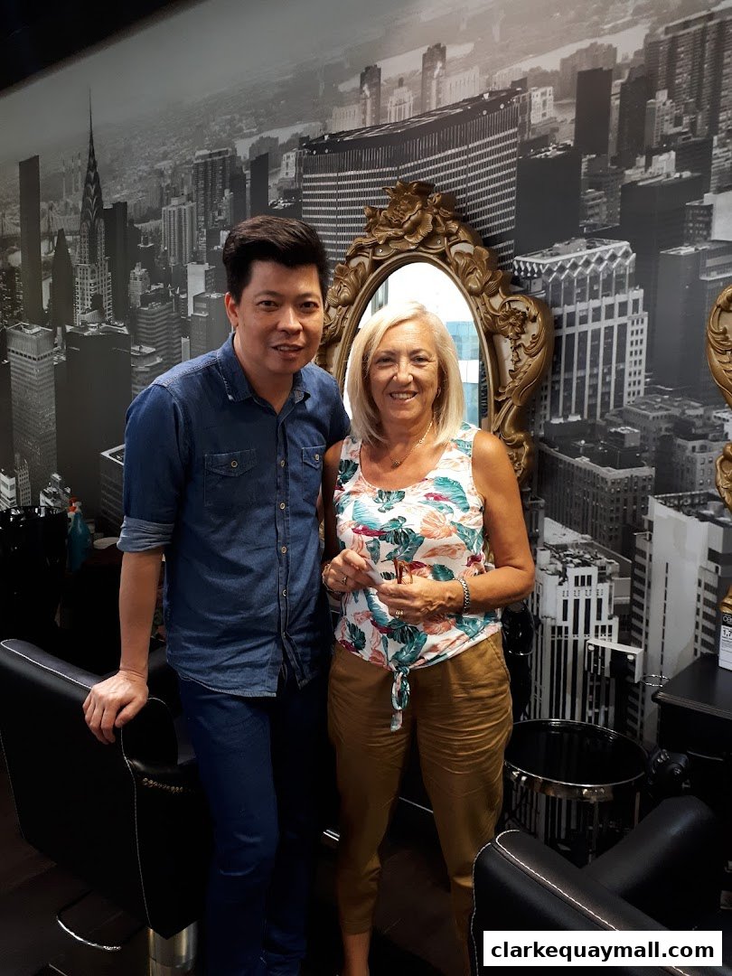 A male stylist and a female client pose in a salon with a city skyline mural.