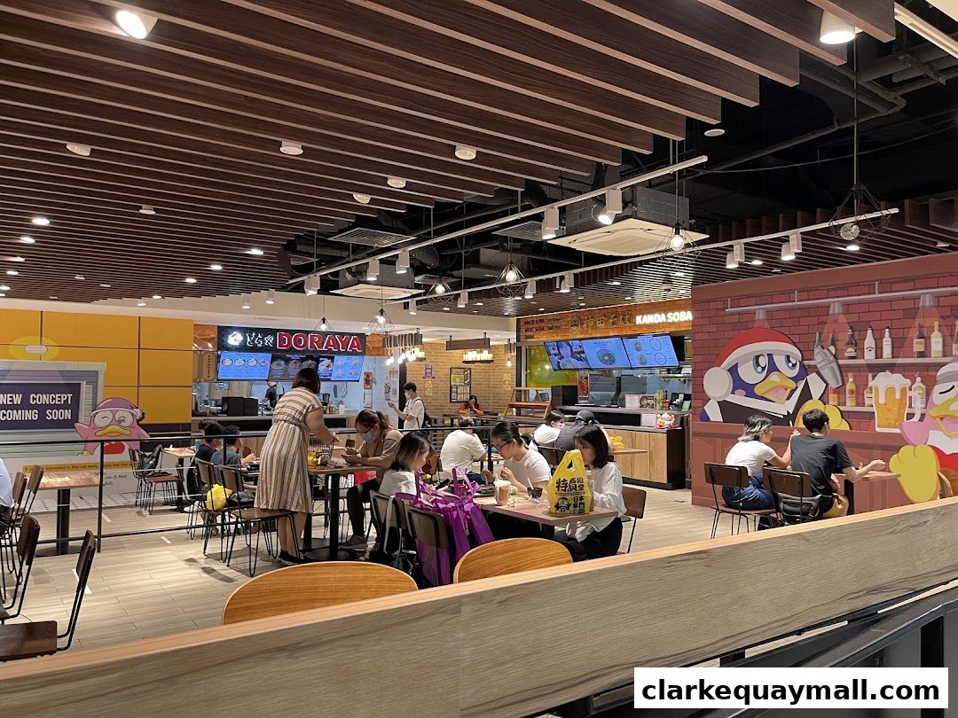 People dining at tables inside a brightly lit food court with various food stalls visible.
