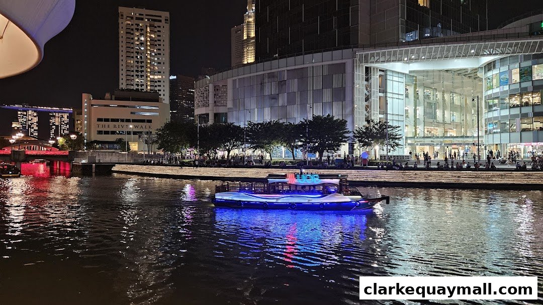 A boat with blue and pink lights sails on a river at night, with a modern building and cityscape in the background.