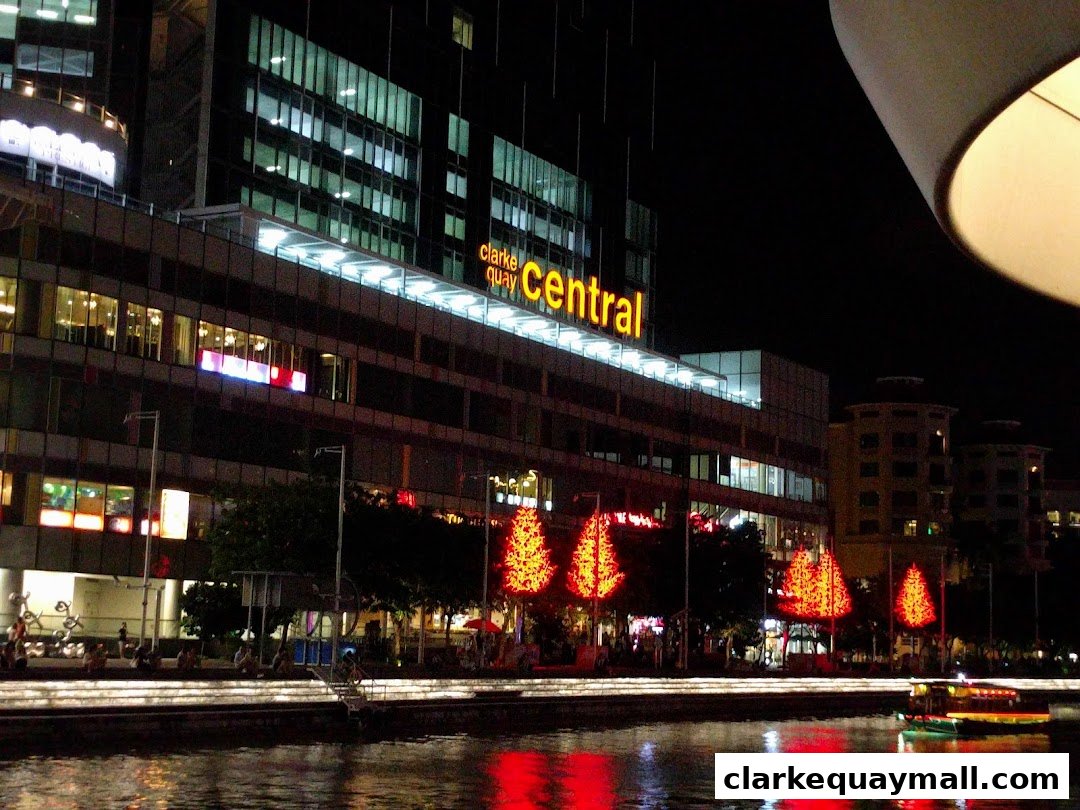 A nighttime view of Clarke Quay Central with illuminated trees and a boat on the river.