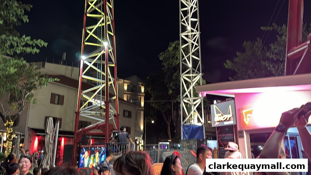 People gather at night around a tall swing ride with bright lights and a neon sign.