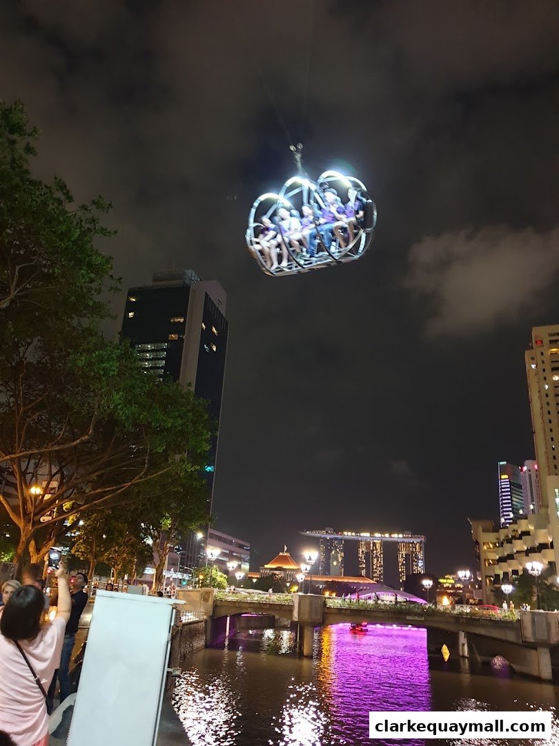 People ride the GX-5 Extreme Swing attraction at night with a city skyline and river in the background.