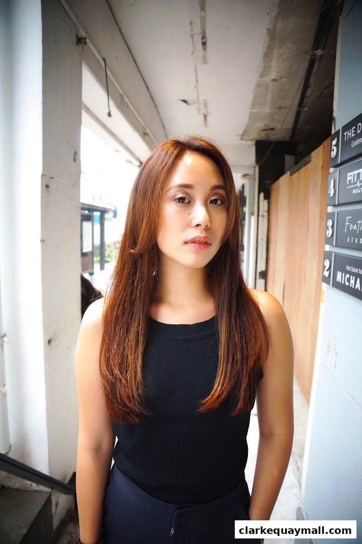 A woman with long, brown hair stands in front of a building with signage for various shops.