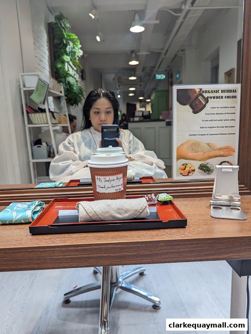 A woman in a salon cape takes a selfie with a coffee cup and a product display in the mirror.