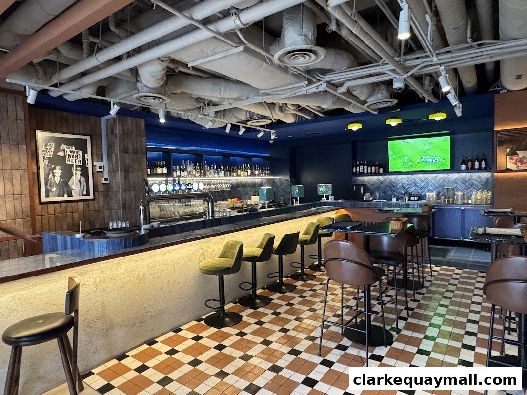 Interior of a bar with a long counter, bar stools, and a TV screen showing a sports game.