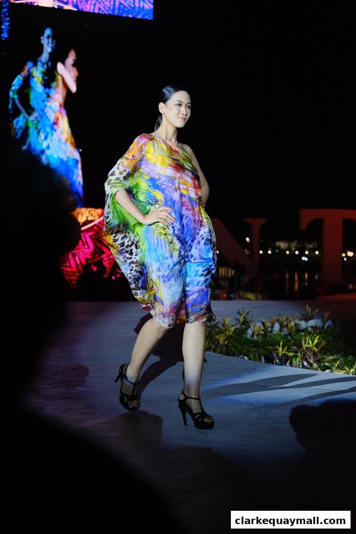 A model walks down a runway wearing a colorful, flowing dress and black heels.