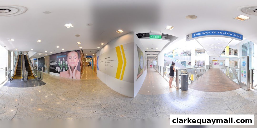 A panoramic view of a modern shopping mall interior with escalators, signage, and shops.