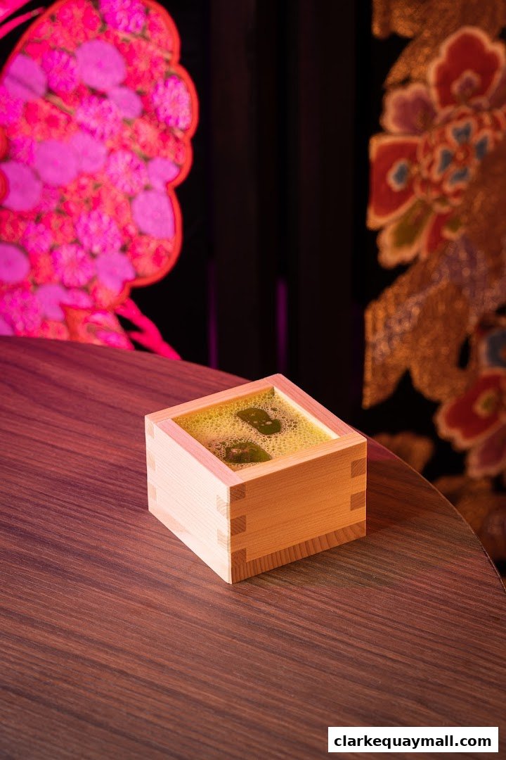 A wooden sake cup filled with a frothy green beverage and garnishes sits on a wooden table.