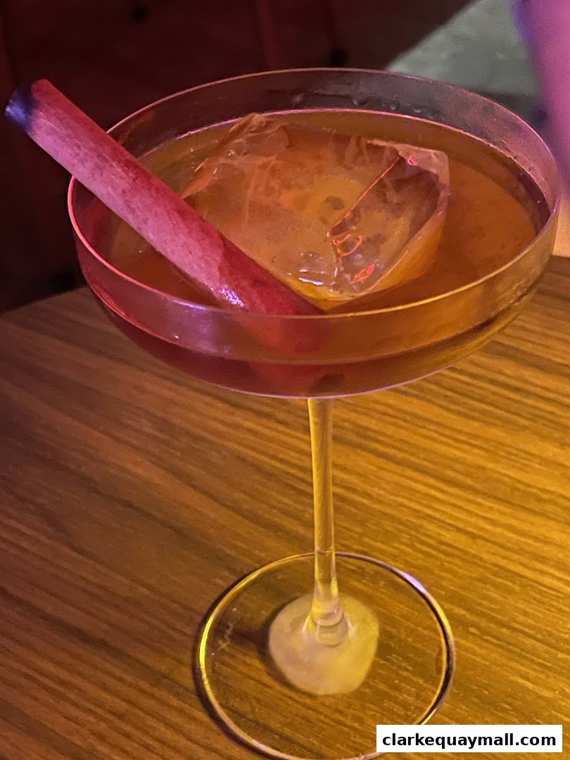 A close-up of a cocktail with a cinnamon stick and large ice cube in a coupe glass.