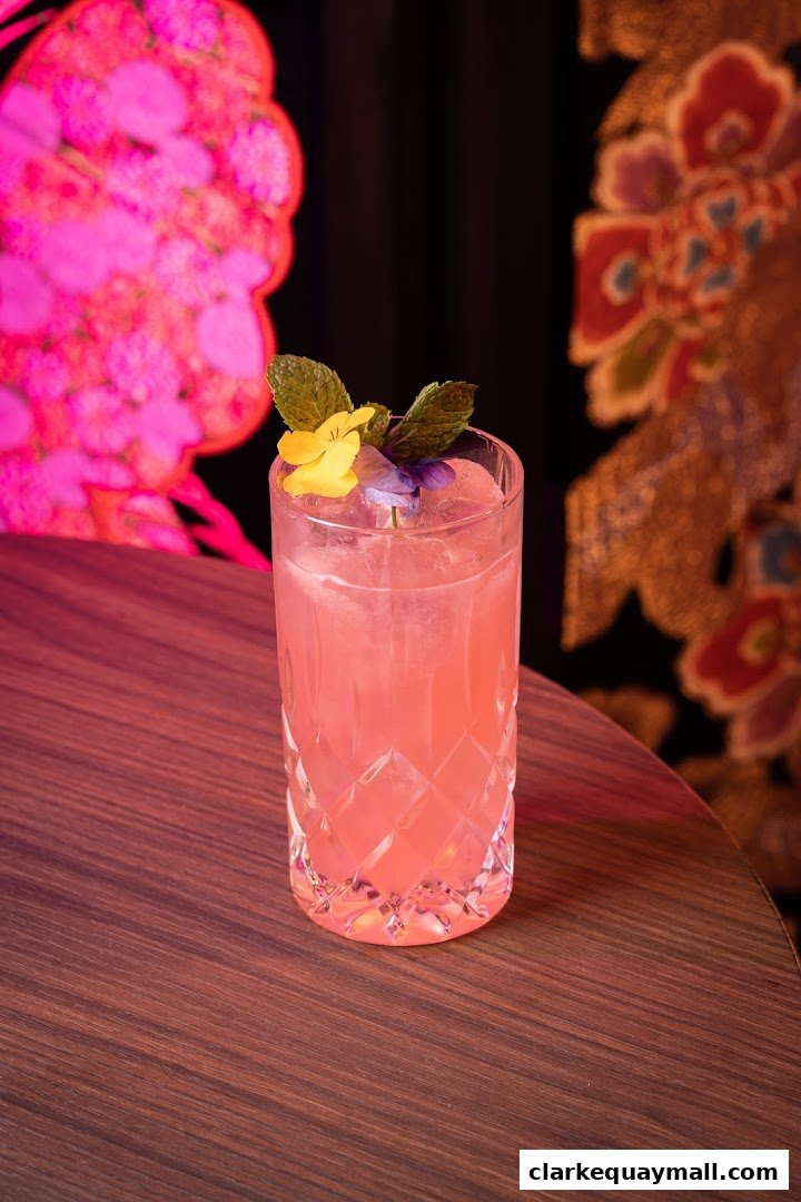 A tall, pink cocktail garnished with mint and edible flowers sits on a wooden table.