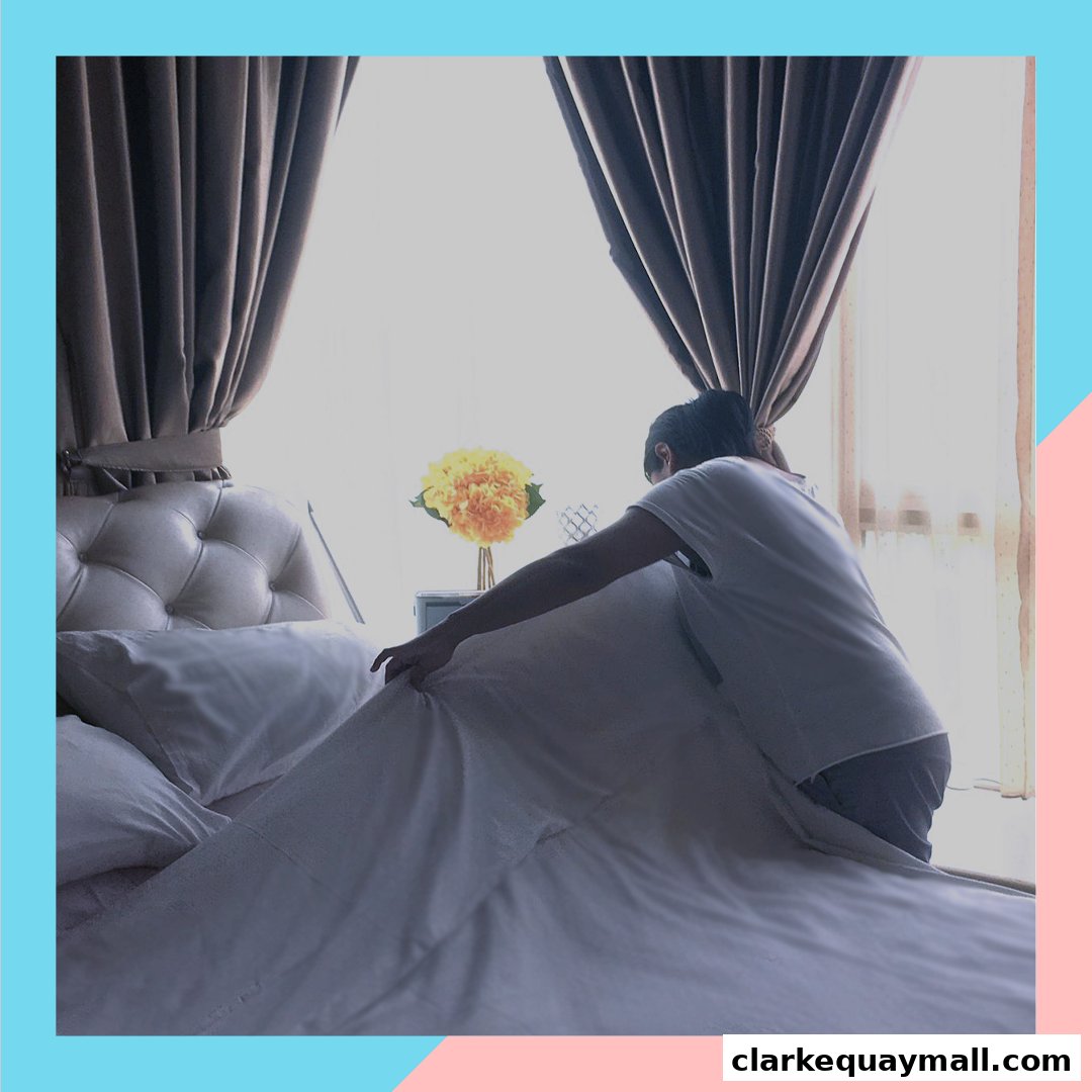 A person making a bed with white sheets and pillows, with curtains and a flower arrangement.