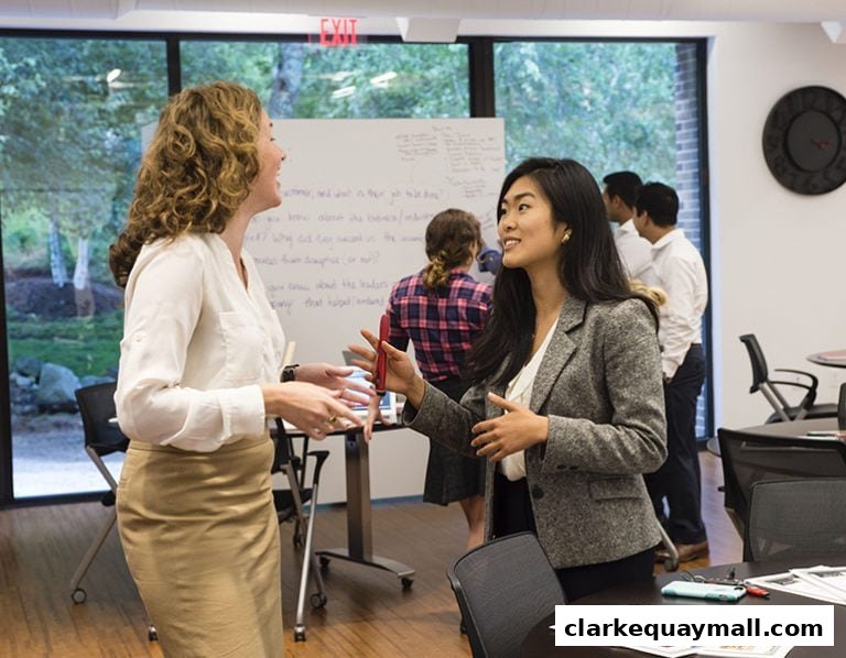 Two women in business attire converse in an office setting with a whiteboard and other colleagues present.
