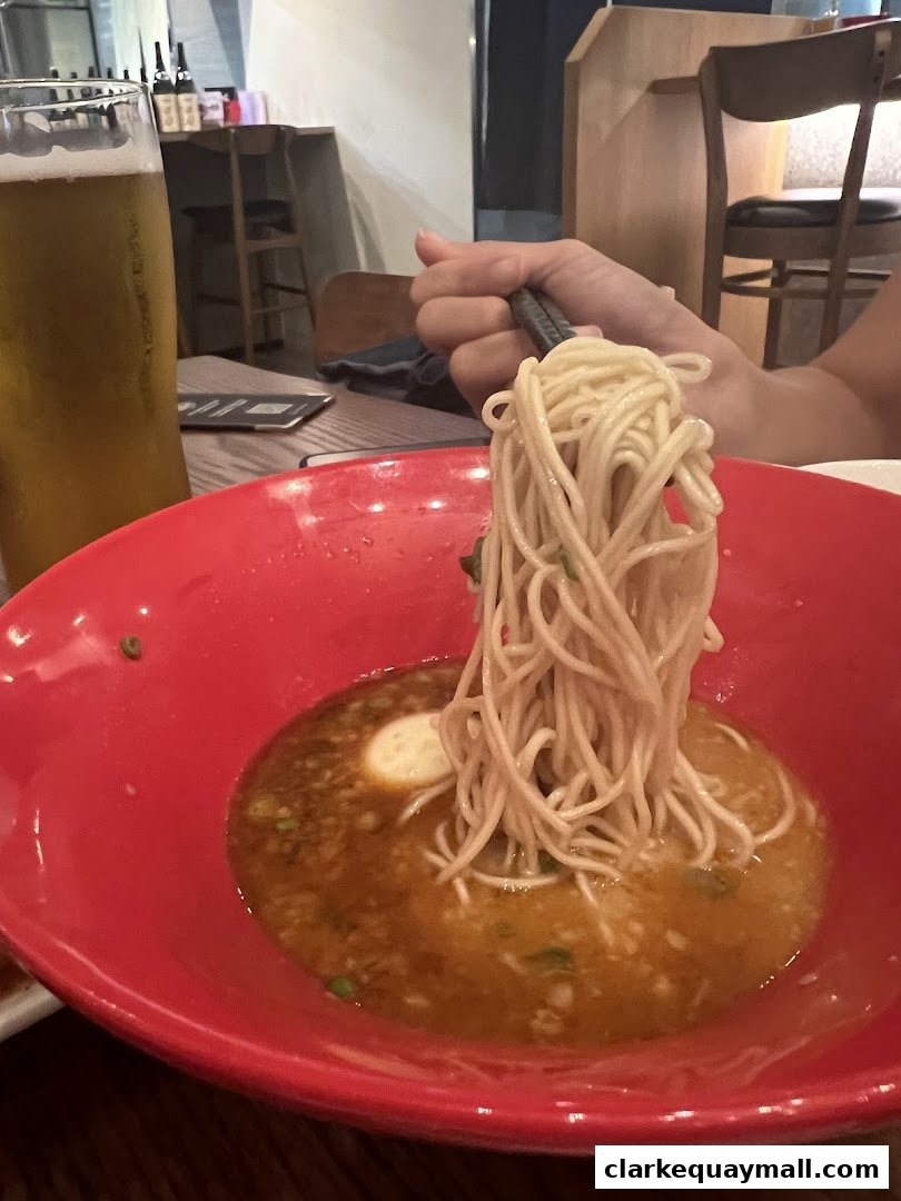 A person is eating ramen with chopsticks from a red bowl, with a glass of beer nearby.