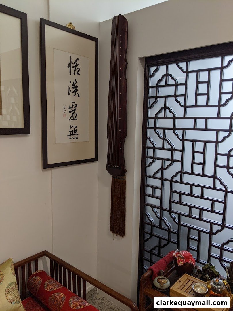 A traditional Chinese guqin instrument hangs on a wall next to calligraphy and a decorative screen.