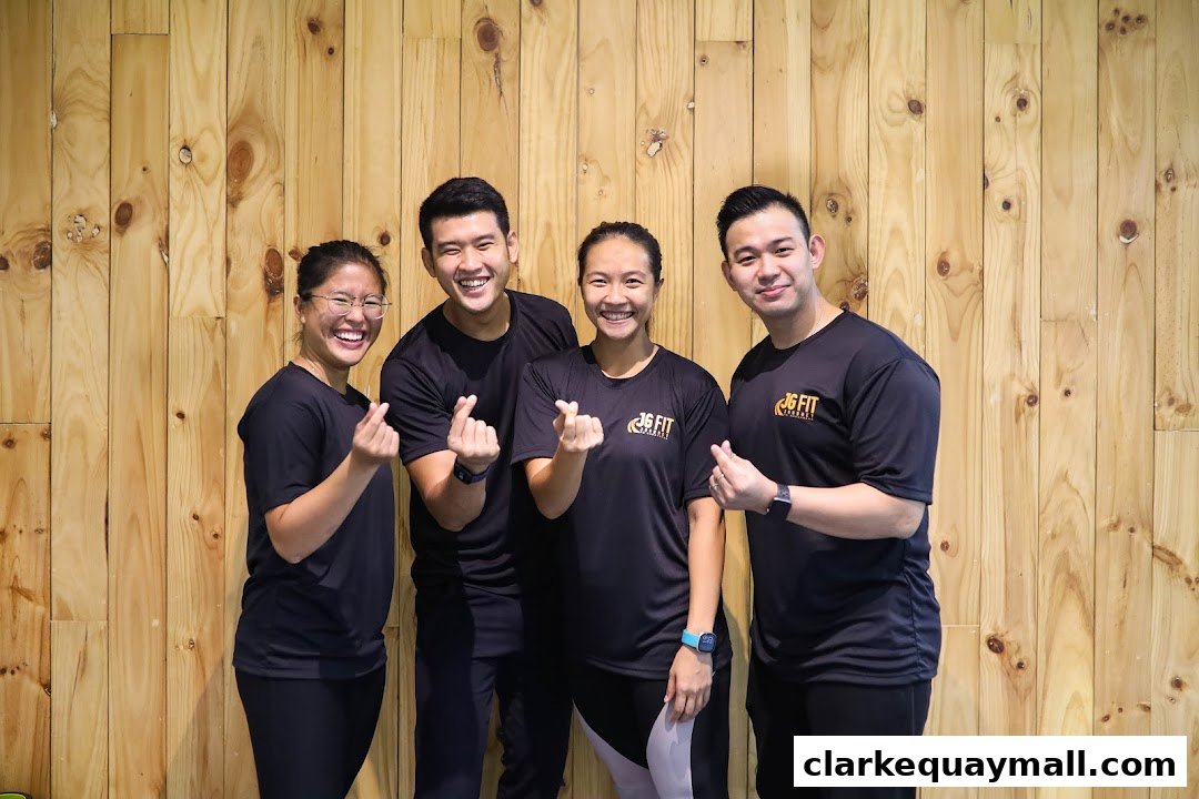 Four smiling people in black JG FIT t-shirts pose against a wooden wall.